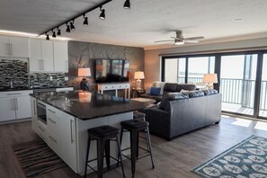 A modern kitchen with a central island, bar stools, a flat-screen TV, and a dining area with a sofa and lamps. - Luxury Fort Myers Beach Penthouse - Stunning Beachfront View & Pool (Fort Myers Beach)