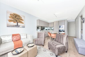 Ein modernes Wohnzimmer mit einer Couch, Sesseln und einem Couchtisch. Es gibt eine Küchenzeile mit einer Mikrowelle und einem Esstisch mit Stühlen.
