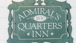 Ein grünes Schild mit dem Schriftzug "Admiral's Quarters Inn 1830" an einer weißen Wand.