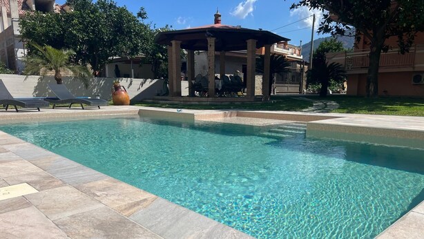 A clear swimming pool with tiled edges, a covered seating area with chairs, and a palm tree in the background. - A beautiful guesthouse where you can relax and enjoy some quality time. (Lanzara)