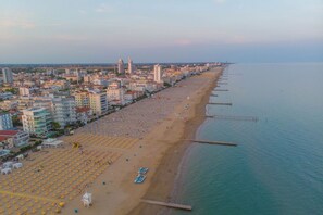 An aerial view of a coastal city with a long beach lined with rows of sunbeds and umbrellas, adjacent to a pier extending into the water. - Villa Nadia 2 - Carraro Immobiliare Jesolo (Lido di Jesolo)