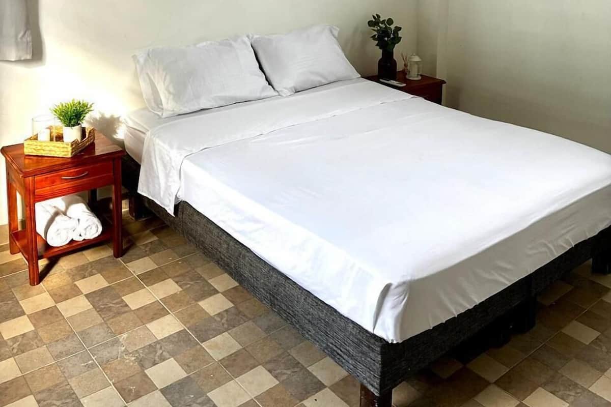 A single bed with white bedding, a wooden nightstand with a plant and a lamp, a ceiling fan, and an air conditioning unit on the wall.