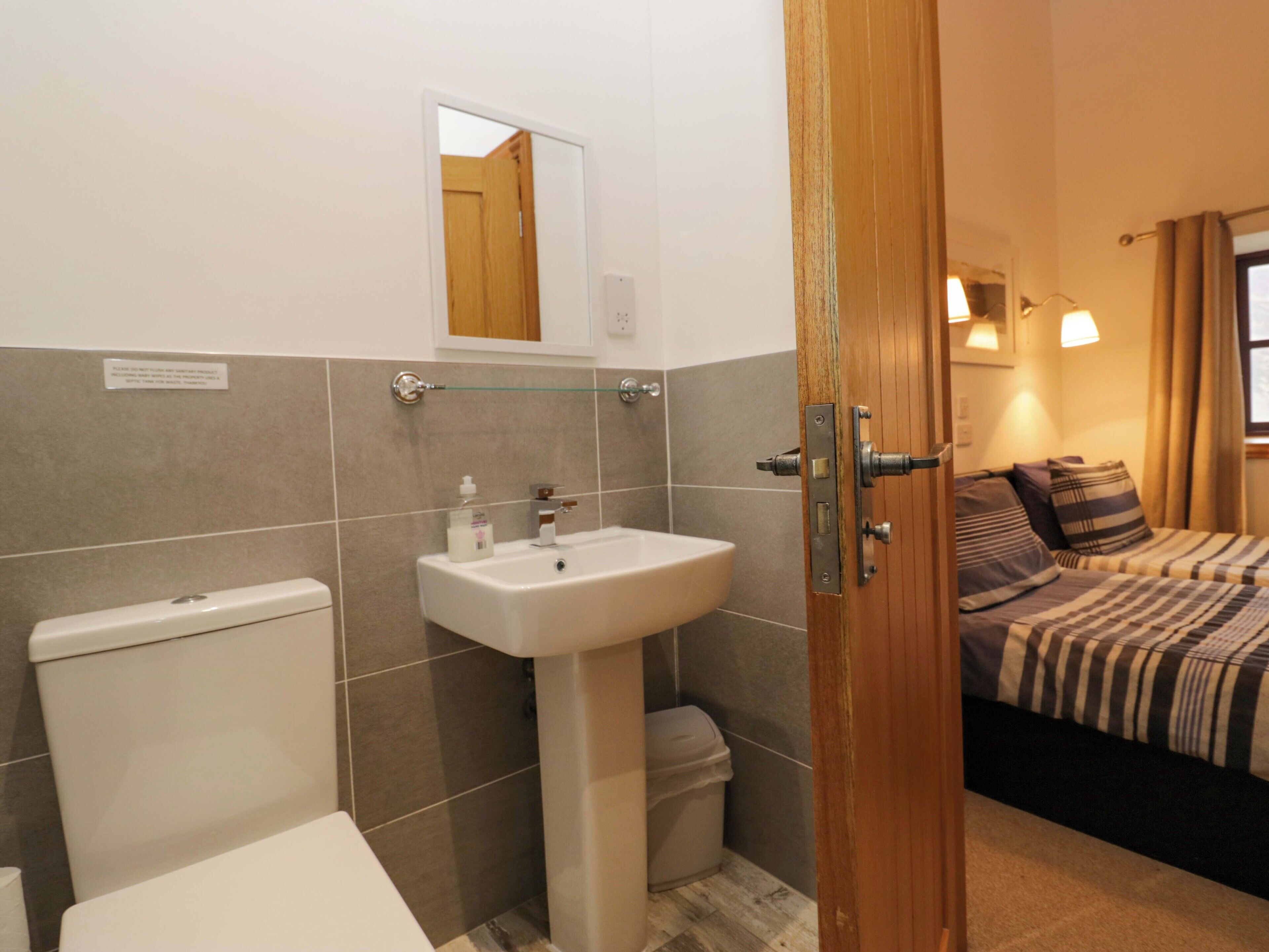 A bathroom with a toilet, sink, and mirror. There is a bed with striped bedding in the adjacent room.