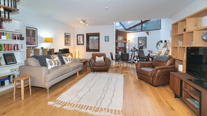 Un salón amplio con un sofá, dos sillones de cuero, una mesa de centro y un mueble para la televisión. Hay estanterías, una zona de comedor y una escalera.