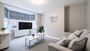 Ein modernes Wohnzimmer mit Flachbildfernseher, rundem Spiegel, grauem Sofa und kleinem Tisch mit Pflanze.