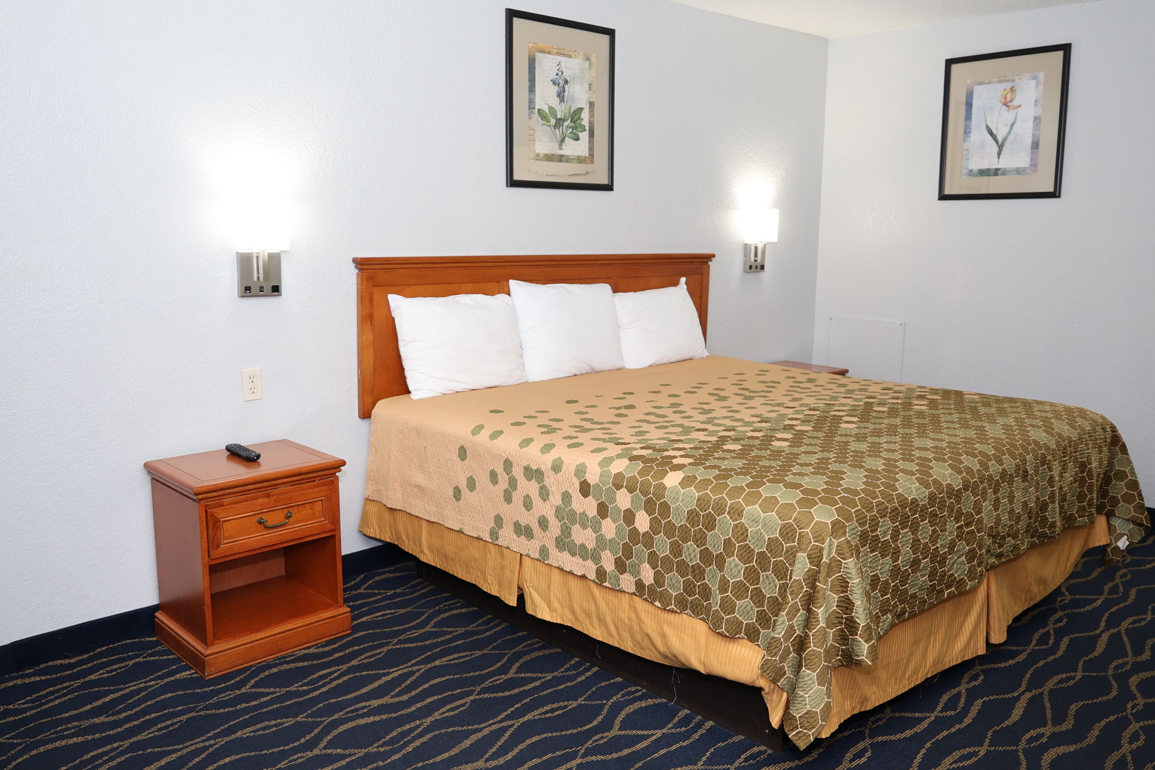 Quarto de hotel com cabeceira de madeira, cama com colcha estampada, mesa de cabeceira com controle remoto e duas luminárias fixadas na parede.