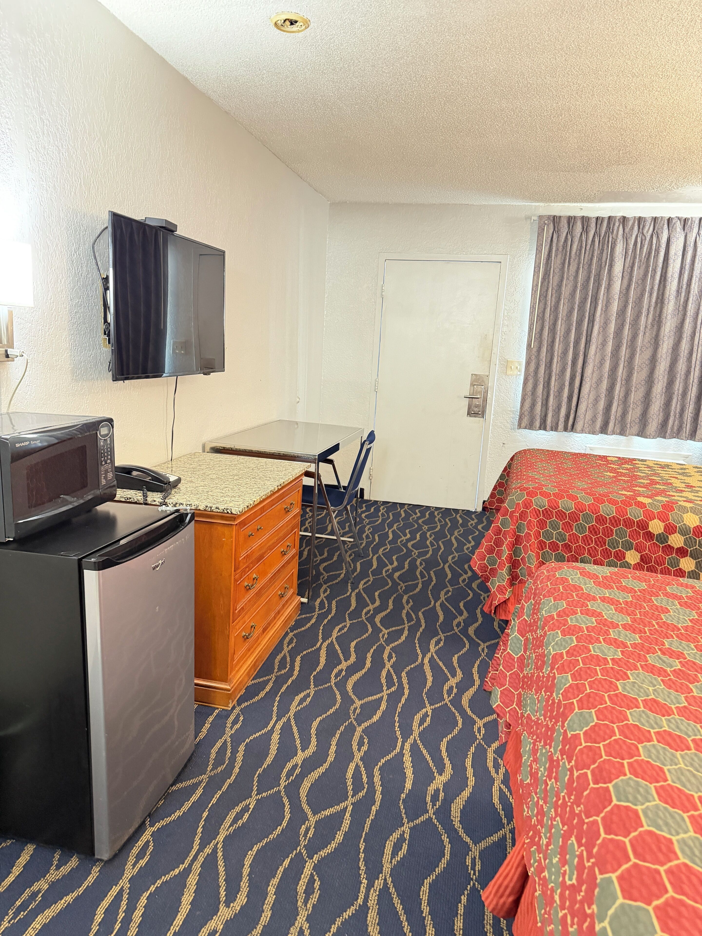 A hotel room with a kitchenette, a bed with a patterned bedspread, a television mounted on the wall, and a door.