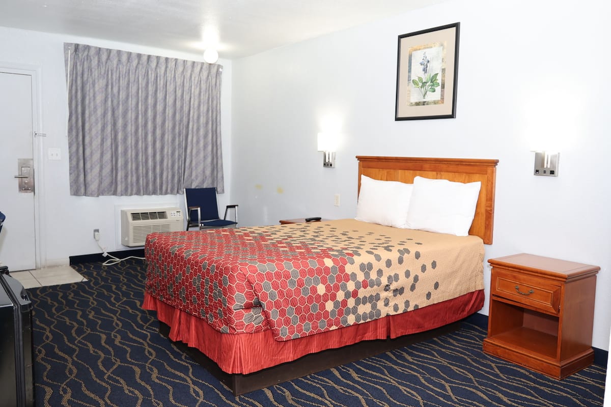 A hotel room with a bed, a desk, an air conditioning unit, a nightstand, and a wall-mounted picture.