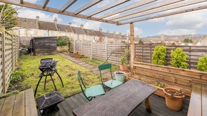 Una pergola in legno sopra un patio con tavolo e sedie, circondato da piante in vaso e vista su un quartiere residenziale.