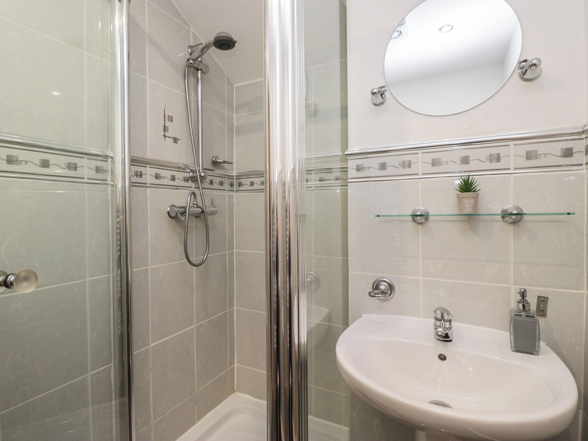 Un bany modern amb plat de dutxa envoltat de vidre, un lavabo blanc amb un mirall damunt i una petita planta en testos sobre una estant.