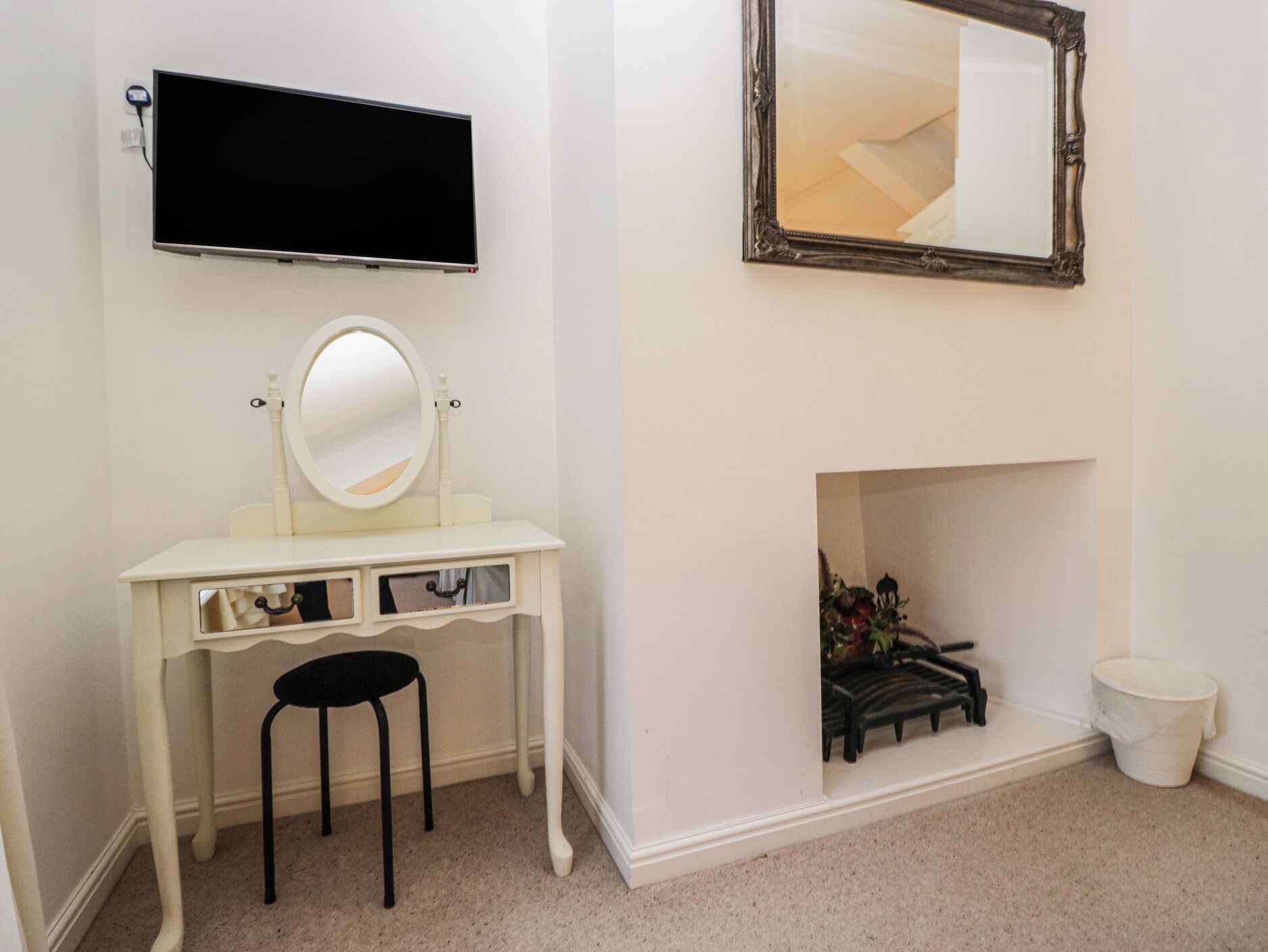 A white vanity with a round mirror and a black stool, a flat-screen TV mounted on the wall, and a fireplace with a decorative mantel.
