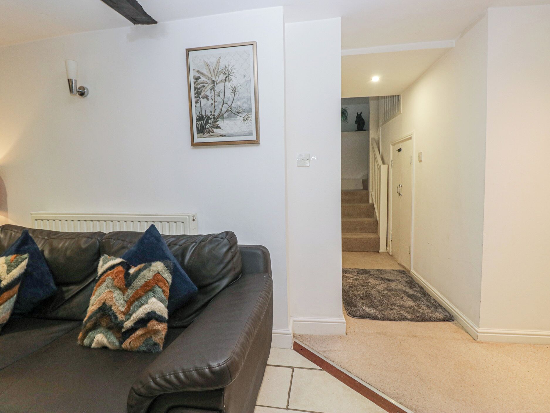A living room with a dark leather sofa, patterned throw pillows, a framed botanical print on the wall, and a carpeted staircase leading to a door.
