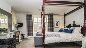 Ein Bett mit Baldachin und Holzrahmen, ein Nachttisch, ein Sessel, ein Schreibtisch mit Spiegel und ein Fenster mit Vorhängen.