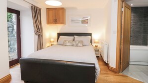 A bedroom with a large bed, wooden cabinets, a ceiling light, and a view of a stone wall and a door. - Newlands Cottage (Keswick)