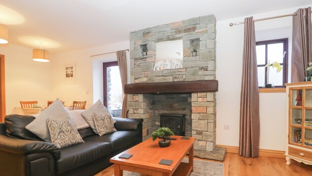 A cozy living room with a stone fireplace, a black leather sofa, and a wooden coffee table. - Newlands Cottage (Keswick)