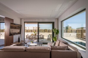 A modern living room with a large window, a sofa, a TV, and a balcony with outdoor furniture. - Villa Sol: Luxury Living in Heart of Gournes (Gournes Gouvon)