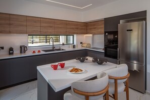 A modern kitchen with a central island, stainless steel appliances, and a large window. - Villa Sol: Luxury Living in Heart of Gournes (Gournes Gouvon)