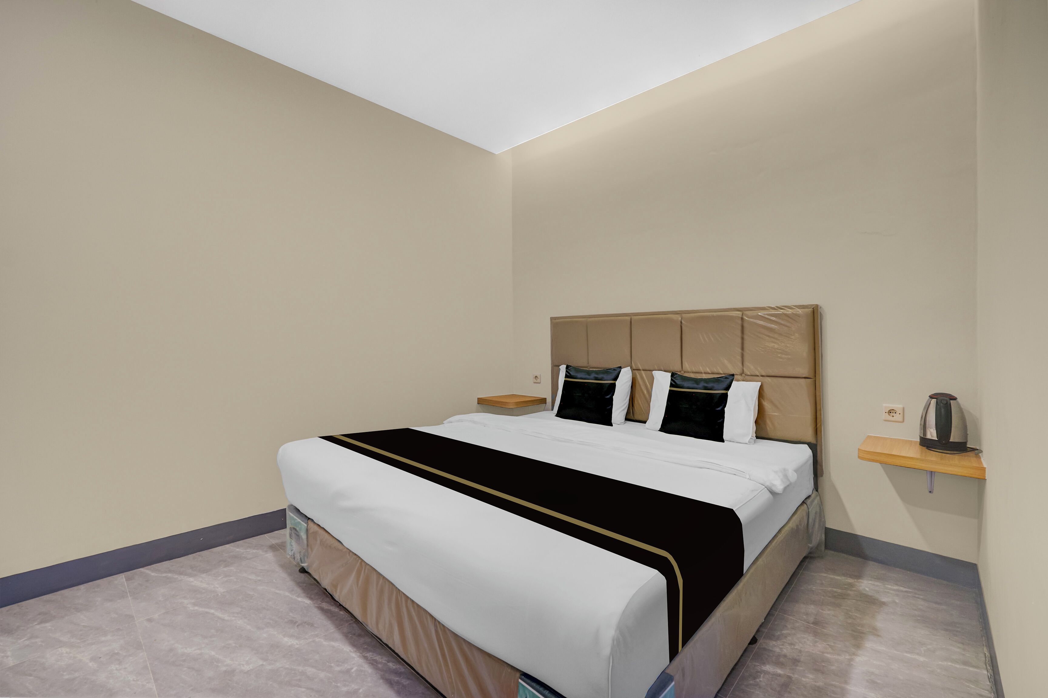A hotel room with a bed, nightstand, and a small shelf with a kettle.