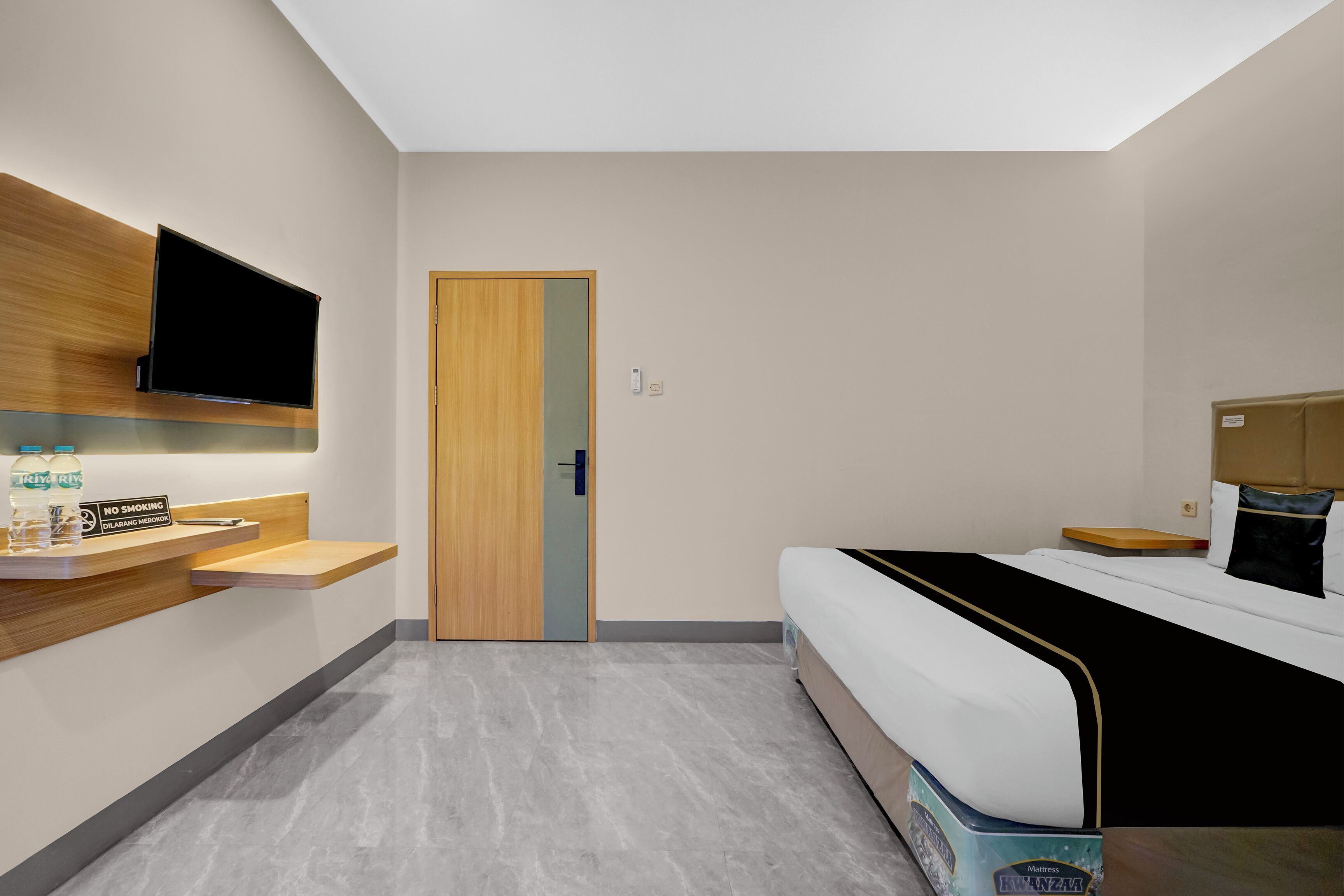 A modern hotel room with a bed, a TV mounted on the wall, a wooden shelf with amenities, and a door.