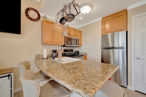 A kitchen with granite countertops, wooden cabinets, stainless steel appliances, and a hanging pot rack. - Waterview 4th Floor Condo | Heated Pool & Hot Tub (St. Petersburg)