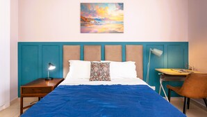 Un dormitorio con cama, escritorio, silla, mesita de noche y un cuadro en la pared.