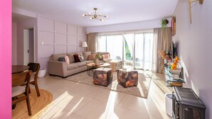Una sala moderna con sofá, una mesa de centro y otomanas, un comedor con sillas y una mesa, y un televisor en la pared.