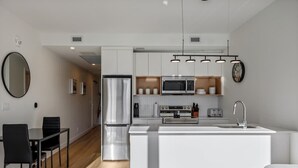 A modern kitchen with white cabinetry, a stainless steel refrigerator, a microwave, and a built-in oven. There is a dining area with a black table and chairs, and a small seating area with a grey sofa. - Sauna, Gym, Pool Table, Walk to Cafes & Sights (Calgary)