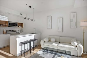 A modern kitchen with white cabinetry, a central island with stools, a white sofa, and framed artwork on the walls. - Gym, Pool Table, Walk to Top Rated Dining, Sauna (Calgary)