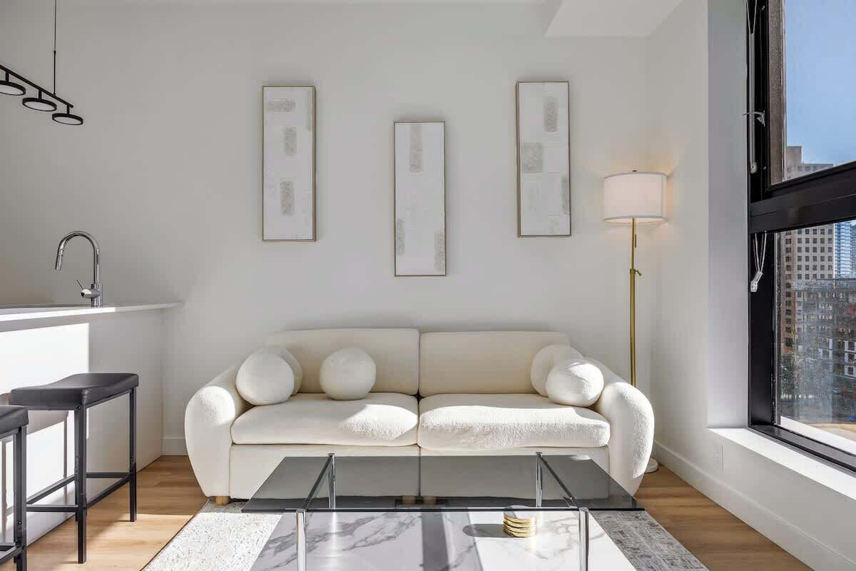 A modern living room with a white sofa, a glass coffee table, and a city view through a large window.