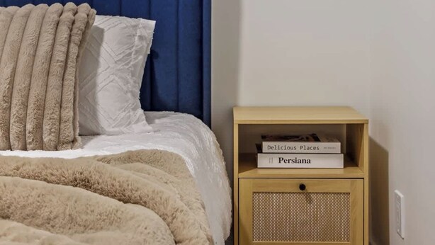 A bedroom with a bed, a blue headboard, a beige blanket, a white pillow, and a wooden nightstand with books. - Steps to Nightlife & Dining, Gym, Sauna, Stampede (Calgary)