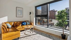 A living room with a sofa, a small table, a floor lamp, a large window with a city view, and a framed artwork on the wall. - Steps to Nightlife & Dining, Gym, Sauna, Stampede (Calgary)