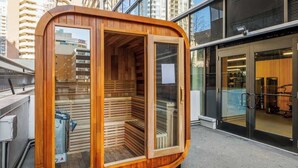 A modern sauna with a wooden interior and glass walls, located in an urban setting with high-rise buildings in the background. - Steps to Nightlife & Dining, Gym, Sauna, Stampede (Calgary)