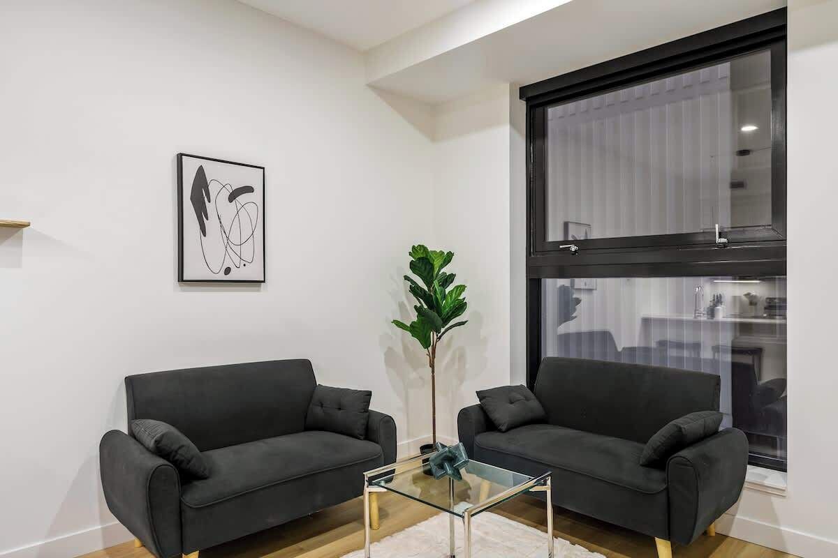 Ein modernes Wohnzimmer mit zwei dunklen Sofas, einem Glastisch, einem großen Fenster und einem minimalistischen abstrakten Gemälde.