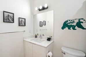 Ein Badezimmer mit weißem Waschbecken, einem Spiegel und einer Toilette. An der Wand befinden sich gerahmte Bilder und ein Bär-Aufkleber.