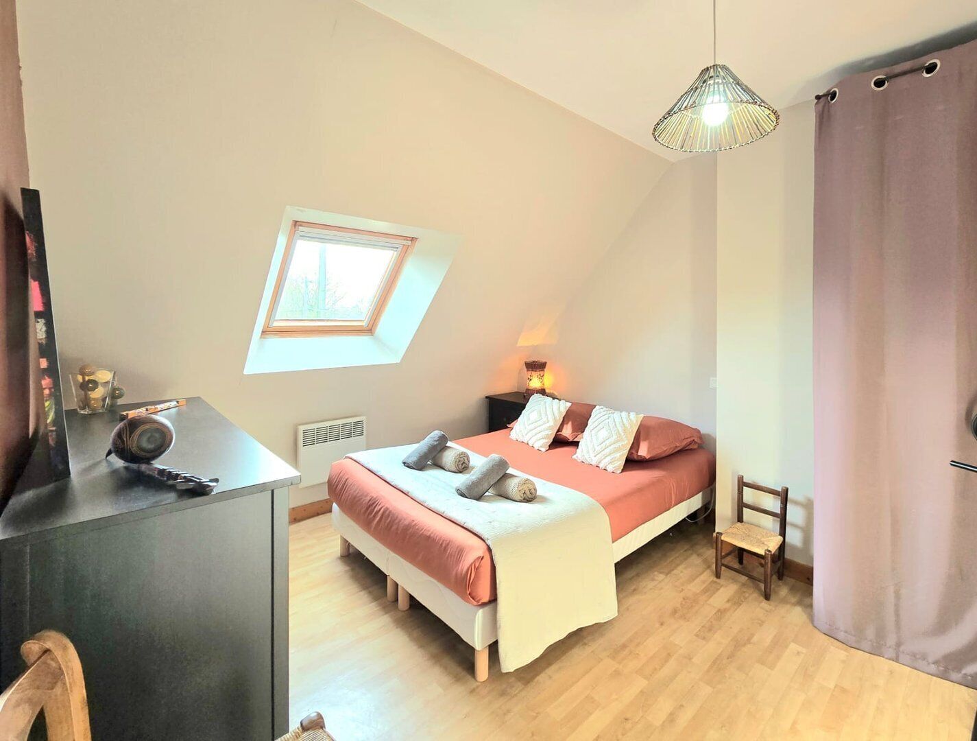 Una camera da letto con soffitto spiovente, un letto con lenzuola rosa, una scrivania con una lampada, una sedia e una finestra vista alberi.