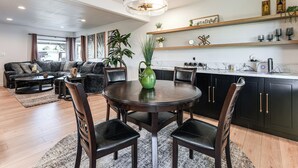 Uma sala de estar moderna com uma mesa de jantar de madeira escura, cadeiras de couro preto e um candelabro. Há um sofá e uma mesa de centro ao fundo.