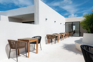 An outdoor seating area with wicker chairs and wooden tables, a white building, and a glass door leading inside. - Elements 326 (Tulum)