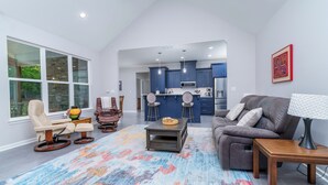 Ein modernes Wohnzimmer mit Sofa, Liege-Sessel, Couchtisch und Lampe. Es gibt eine Küchenzeile mit blauen Schränken und einer Mikrowelle.