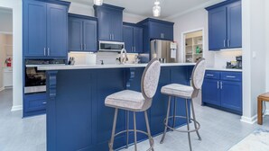 Eine moderne Küche mit blauen Schränken, einer zentralen Kochinsel mit Barhocker, Edelstahlgeräten und Pendelleuchten.