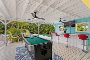 A pool table in a covered outdoor area with a ceiling fan, a bar area with red stools, and a TV screen. - Private Pool, Bar, Putt Green, Jet Tub, King Bed, quiet town 5min drive to beach (Pompano Beach)