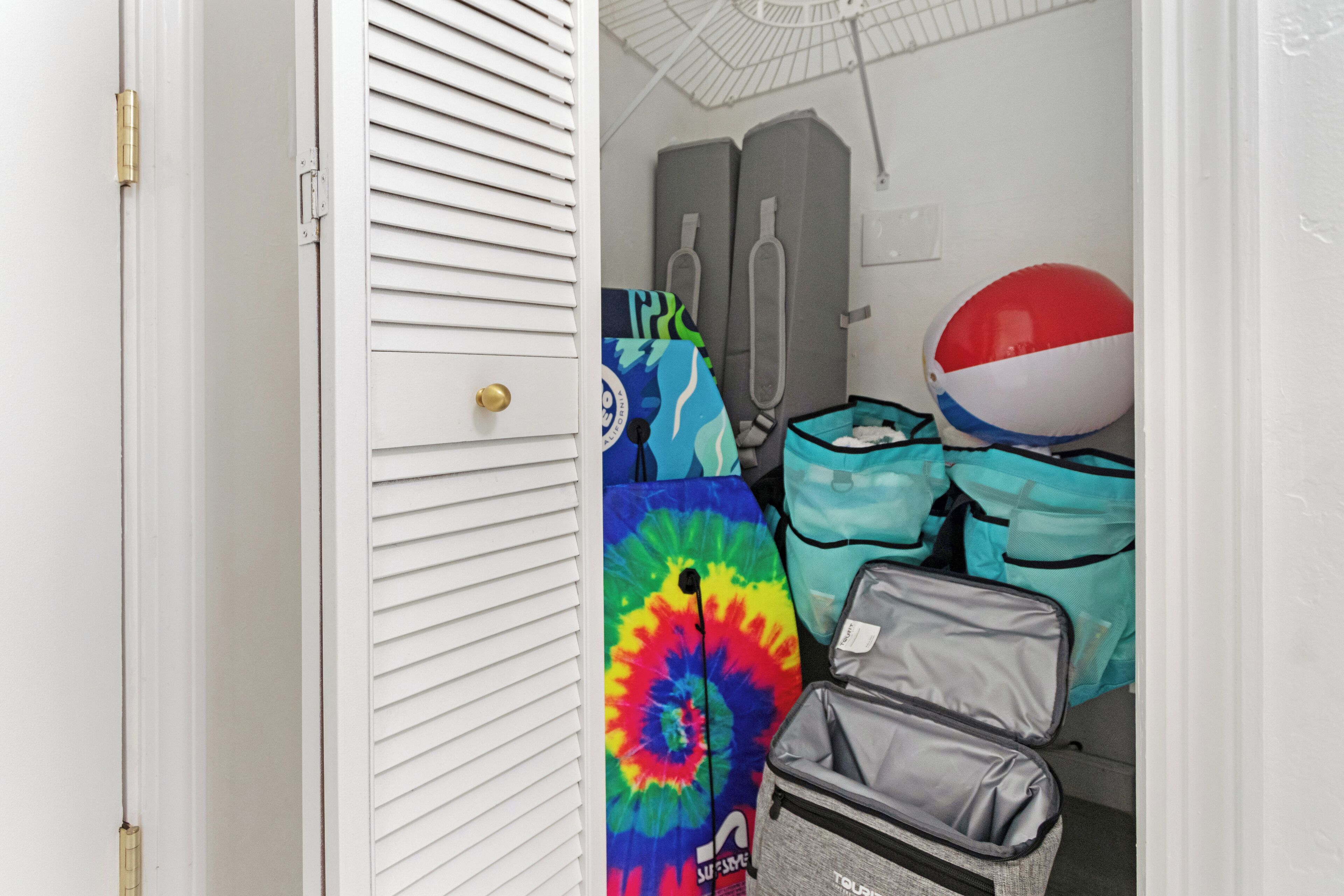 A storage area with a white door, a tie-dye bag, a gray suitcase, and a red and white beach ball.