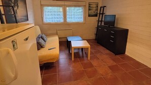 A compact living space with a sofa, a small table, a refrigerator, a TV, and a window with a view of trees.