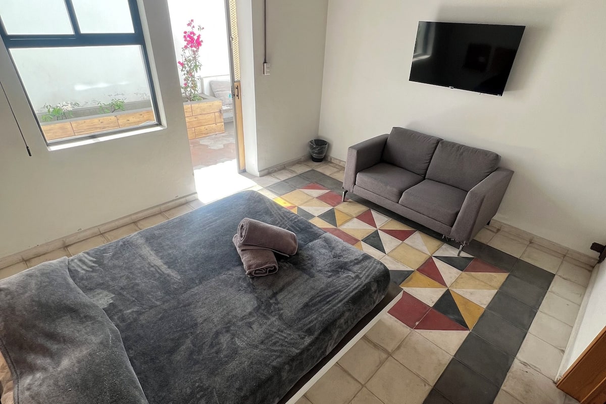 A room with a bed, a sofa, a TV mounted on the wall, and a patterned floor.