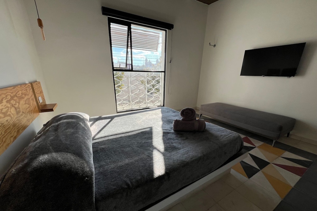 A room with a bed, a bench, a TV mounted on the wall, and a window with blinds.