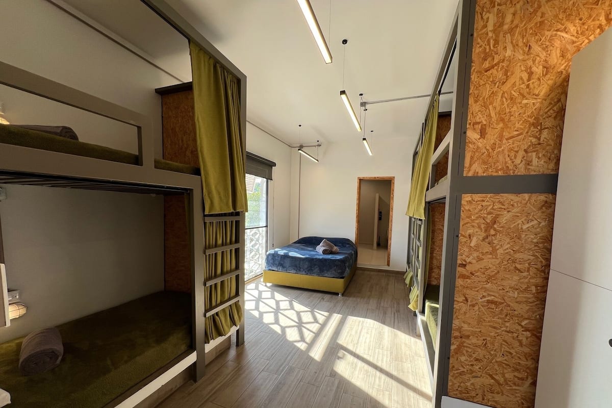 A modern, well-lit room with bunk beds, a single bed, and a large window allowing natural light.