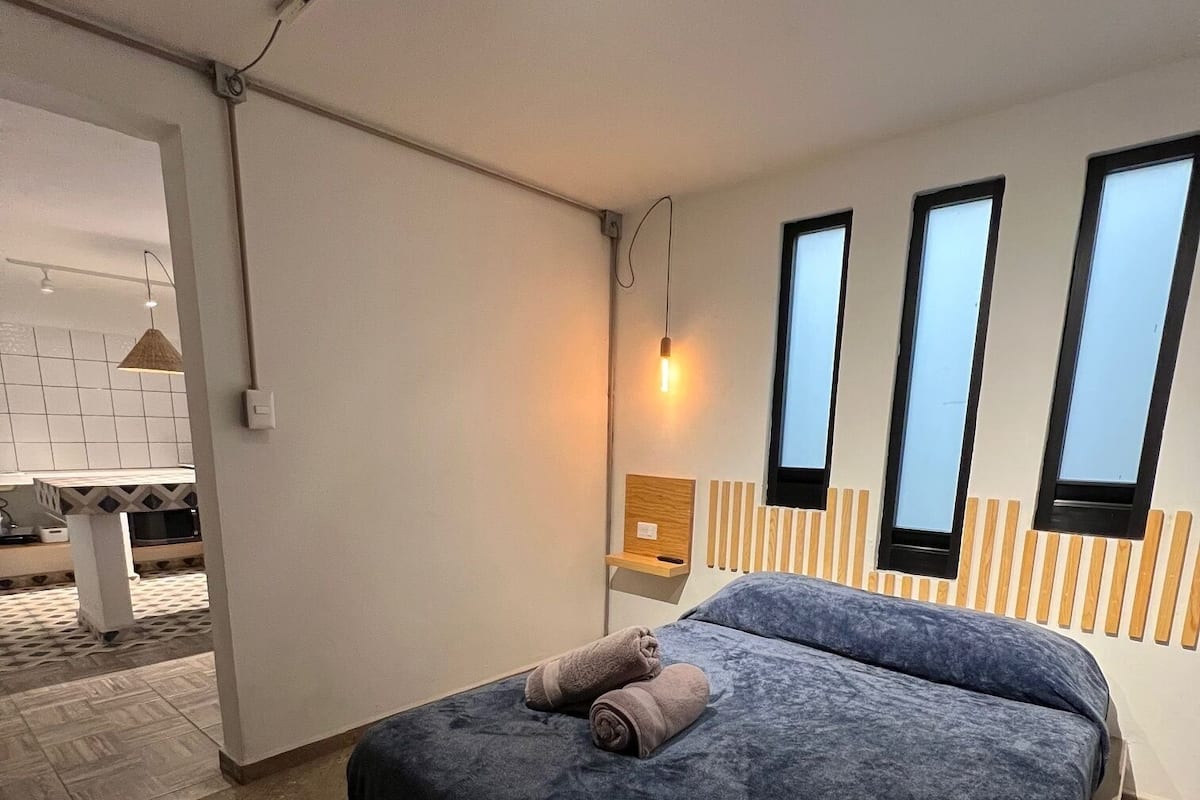 A modern bedroom with a large bed, a wooden headboard, and a small bedside table. There are three windows on the wall, and a hanging light fixture above the bed.