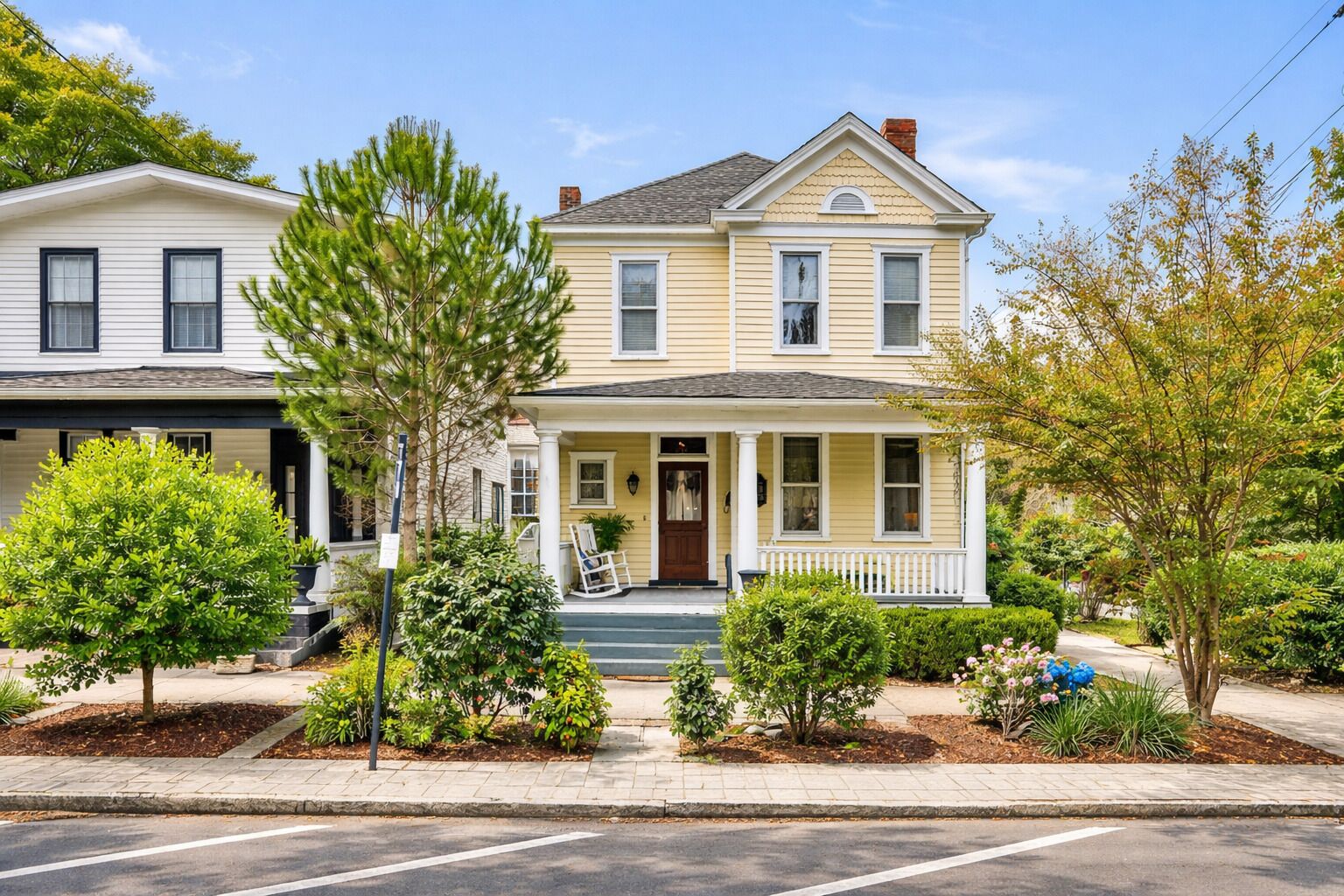 A two-story house with a front porch, a small front yard with trees and shrubs, and a clear sky.