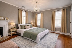Un dormitorio con cama, dos mesitas de noche, un candelabro, chimenea y tres ventanas con cortinas.