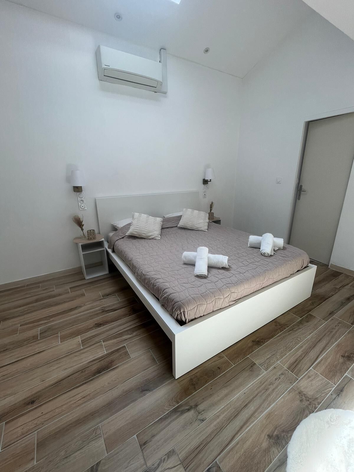 A modern bedroom with a bed, bedside lamps, an air conditioning unit, and a wooden floor.