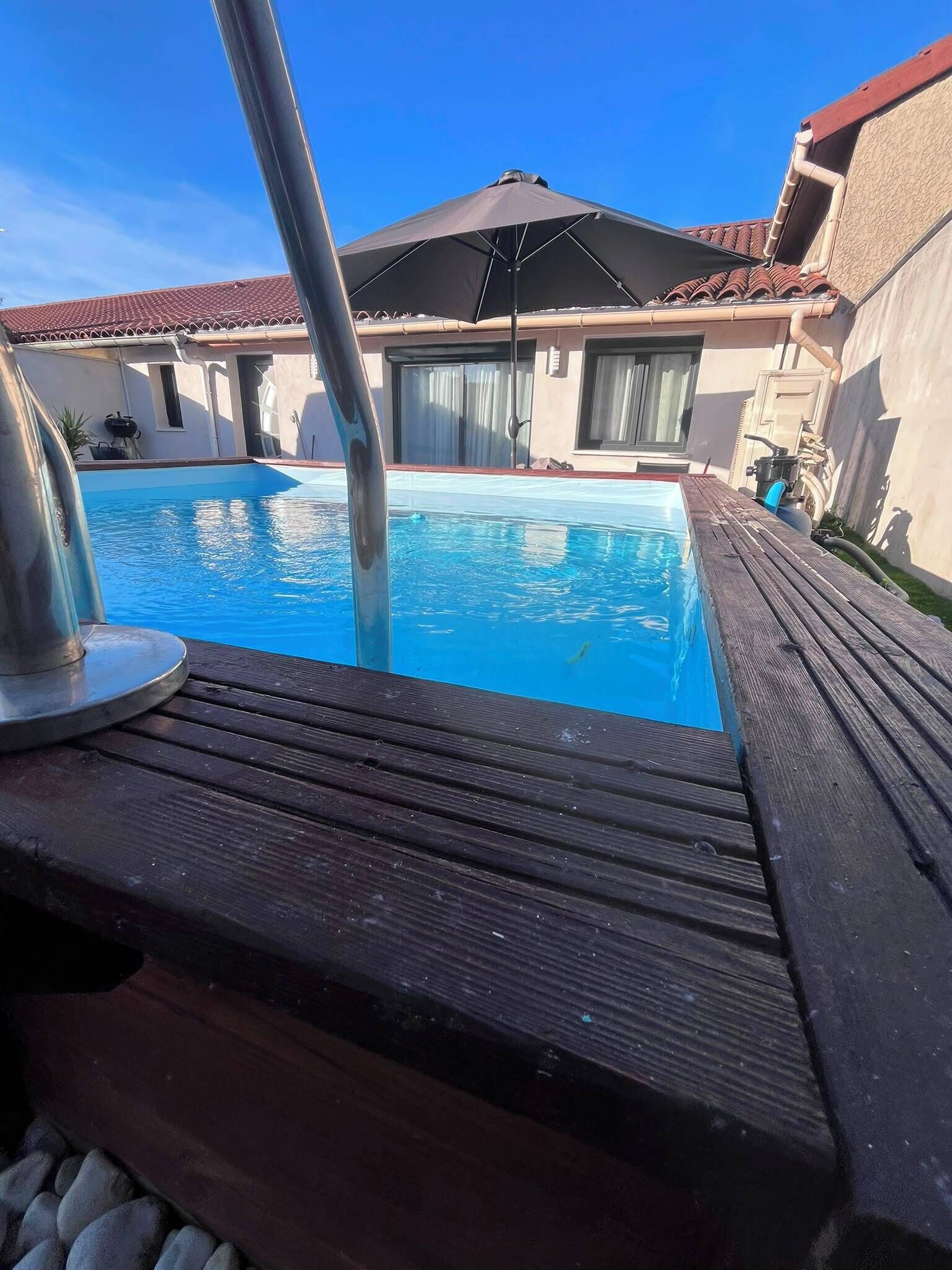 A swimming pool with a wooden deck, a metal ladder, and a covered area with a red-tiled roof.